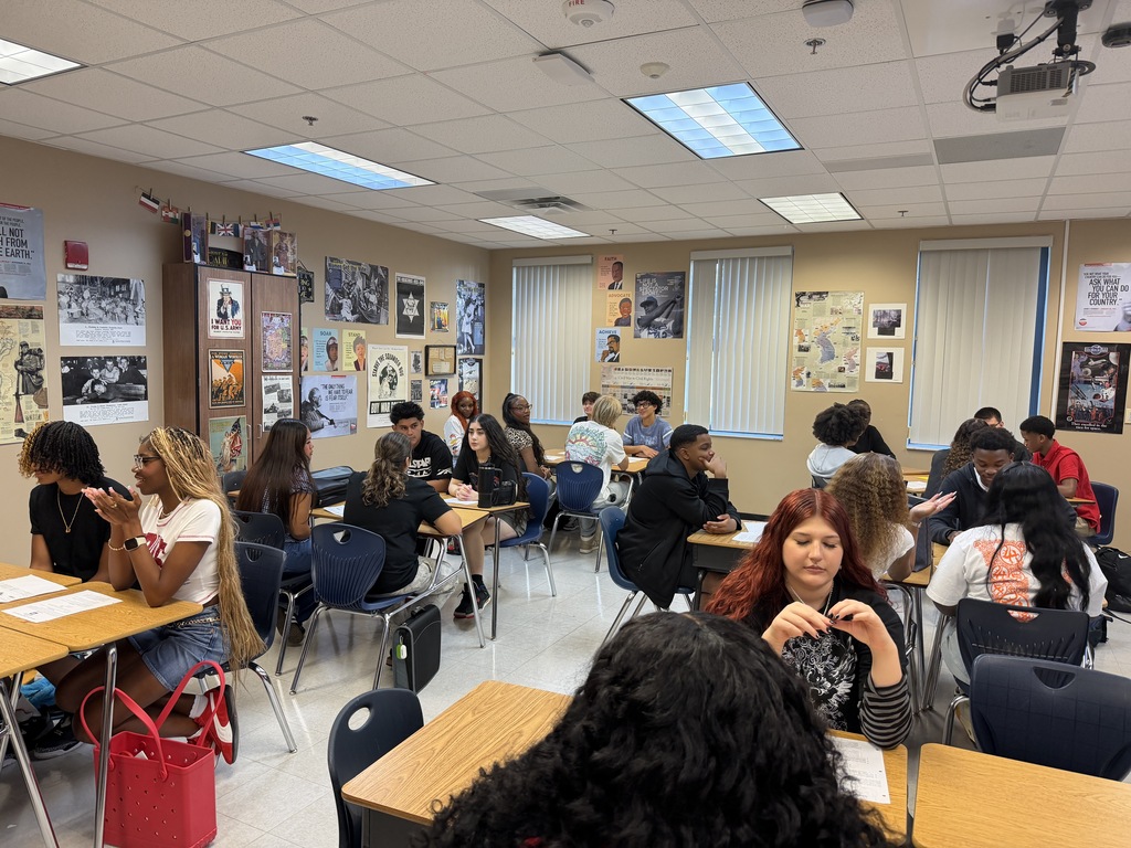 A classroom with people seated at tables. The room has posters on the walls, ceiling lights, and windows with blinds.