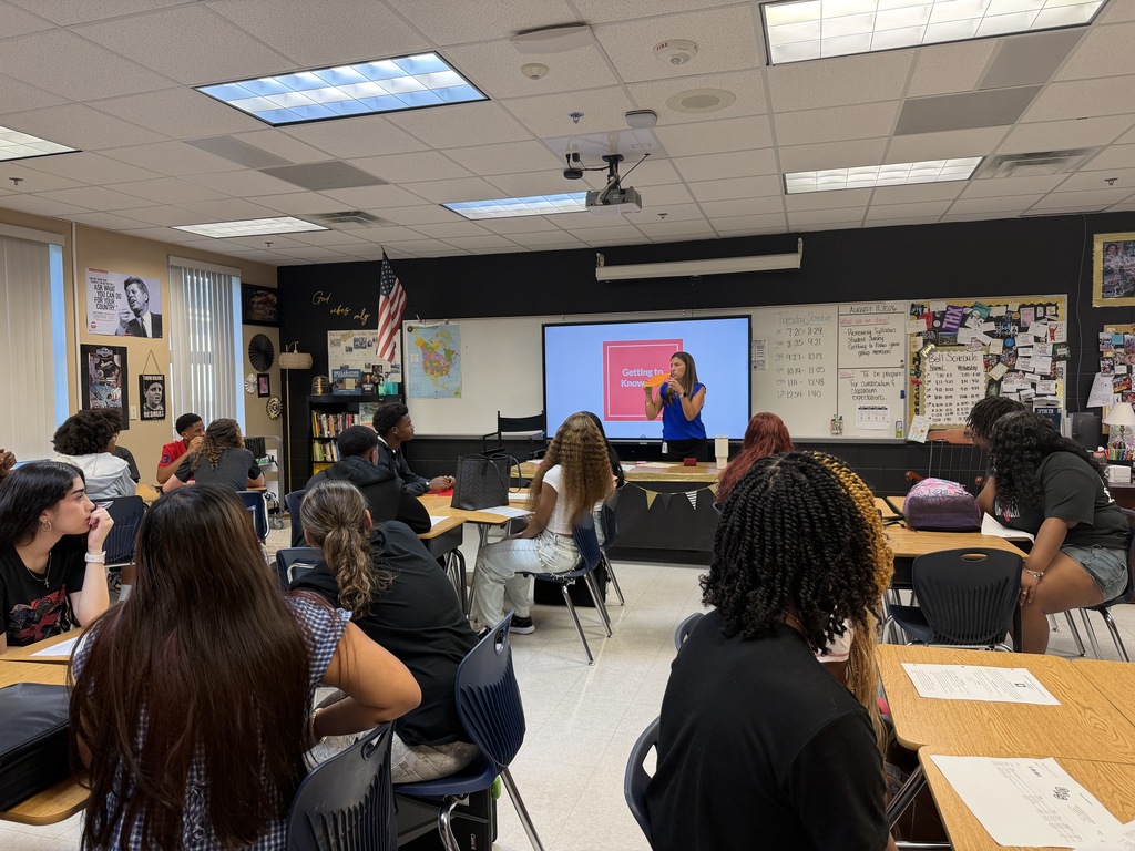 A classroom with students seated at tables. A teacher stands in front of a screen, presenting. The room has ceiling lights and framed pictures on the wall.