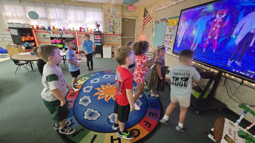 A classroom with children gathered around a monitor, watching and dancing to a video game.