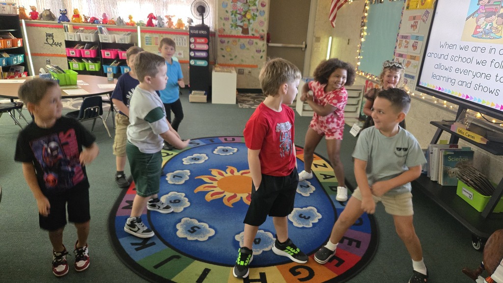 Children stand in a circle on a colorful rug, facing a screen displaying a presentation.