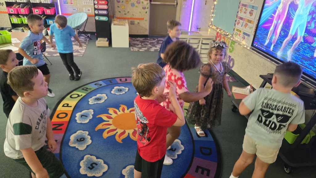 Children in a classroom circle, some wearing t-shirts, gather around a large circular rug with sun design.