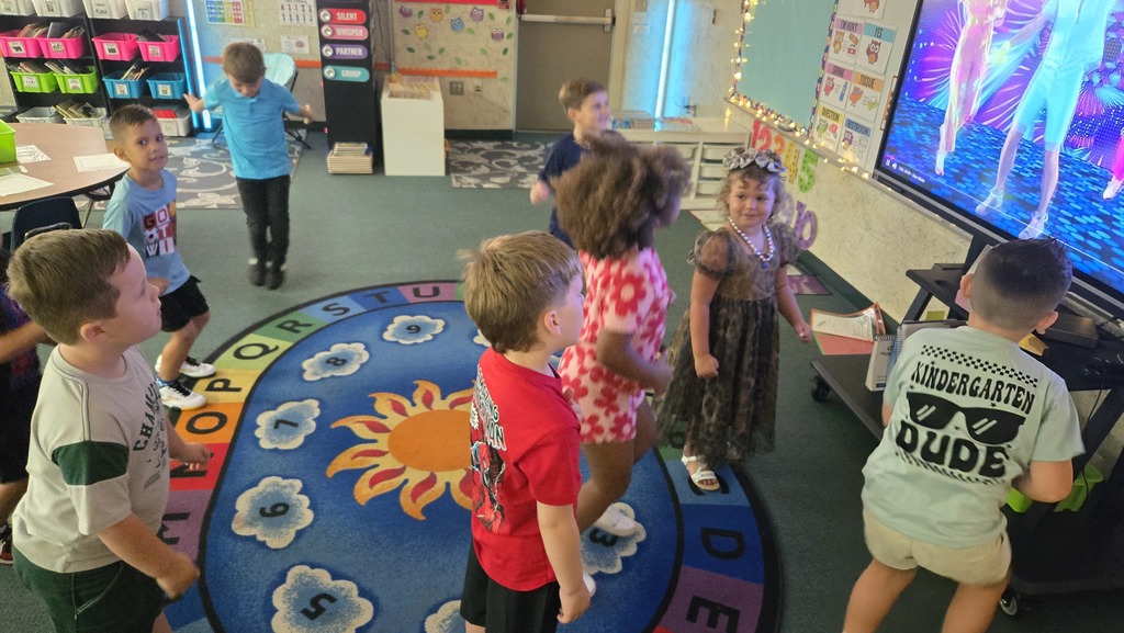 Children stand around a circle rug with letters, some watching a video on a large screen.