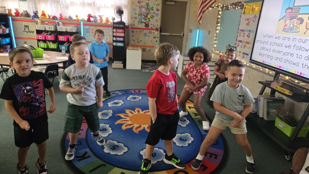 Children in a classroom, some standing and dancing, others sitting, on a multicolored circle rug.