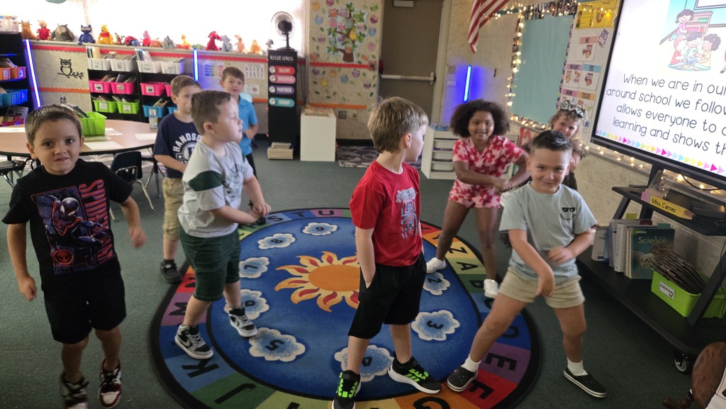Kids in a classroom standing on a multicolored mat, dancing, and a large monitor in the background.