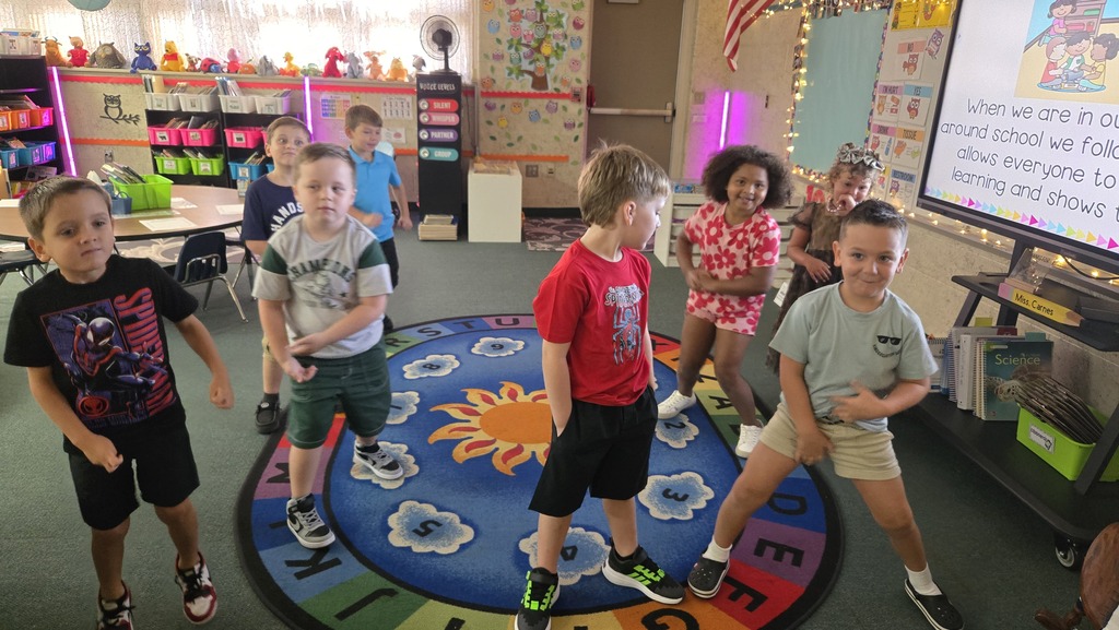 Children in a classroom with a rug. One child stands on a yellow sun, others in front of a screen.