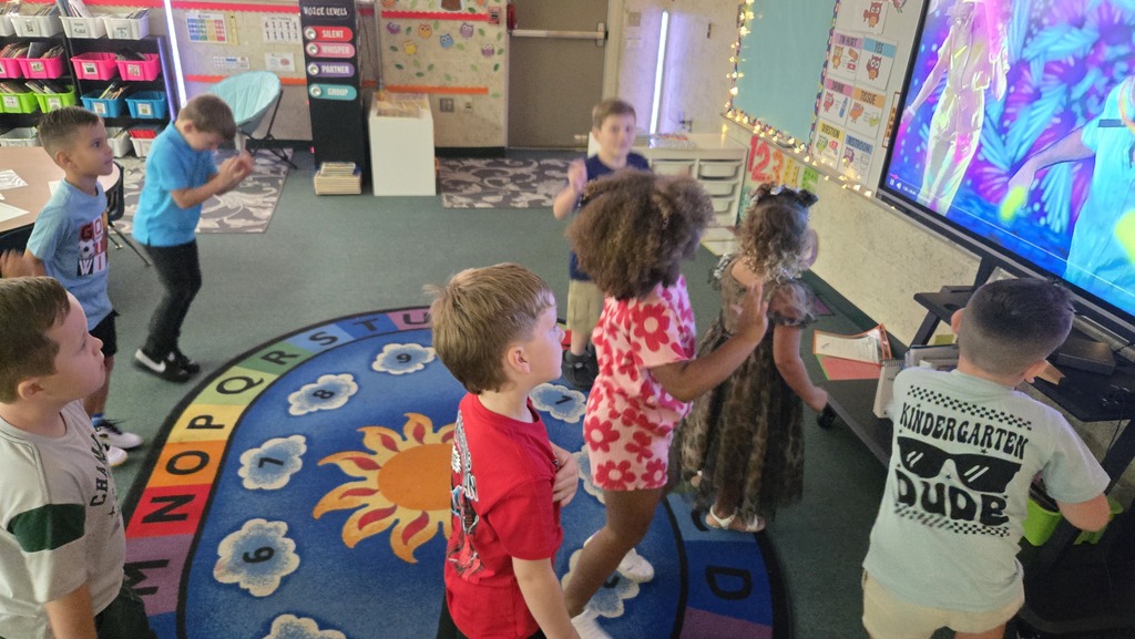 Kids in a classroom with a colorful rug, standing around a large TV playing an educational video.