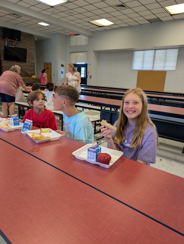 Several children seated at a long table eat food. A woman stands behind them in a cafeteria.
