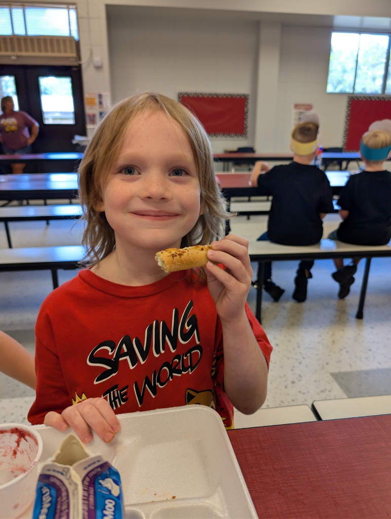 A child wearing a red shirt eats a roll with a bite taken out. Behind them are two people sitting at a table.