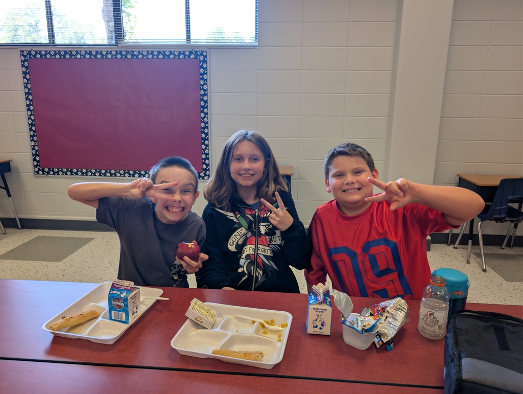 Three children seated at a table with various food items in front of them in a classroom.