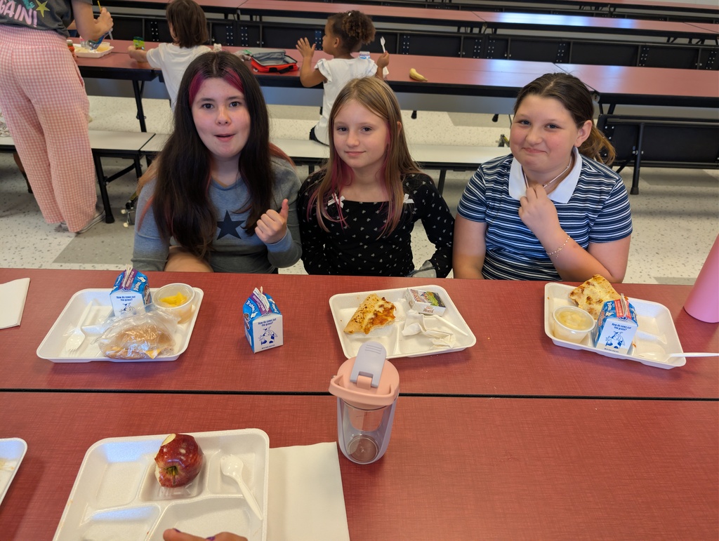 Three girls sit at a cafeteria table with trays of food, one with a thumbs-up.