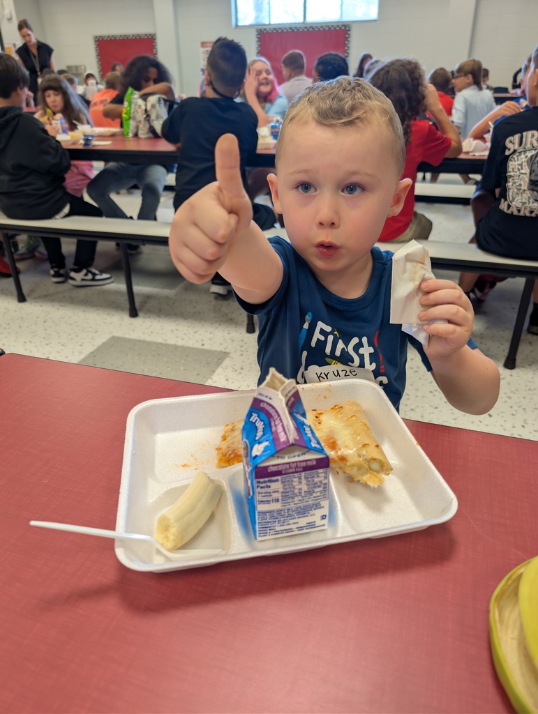 A young boy gives a thumbs up, seated at a cafeteria table with a meal tray before him.