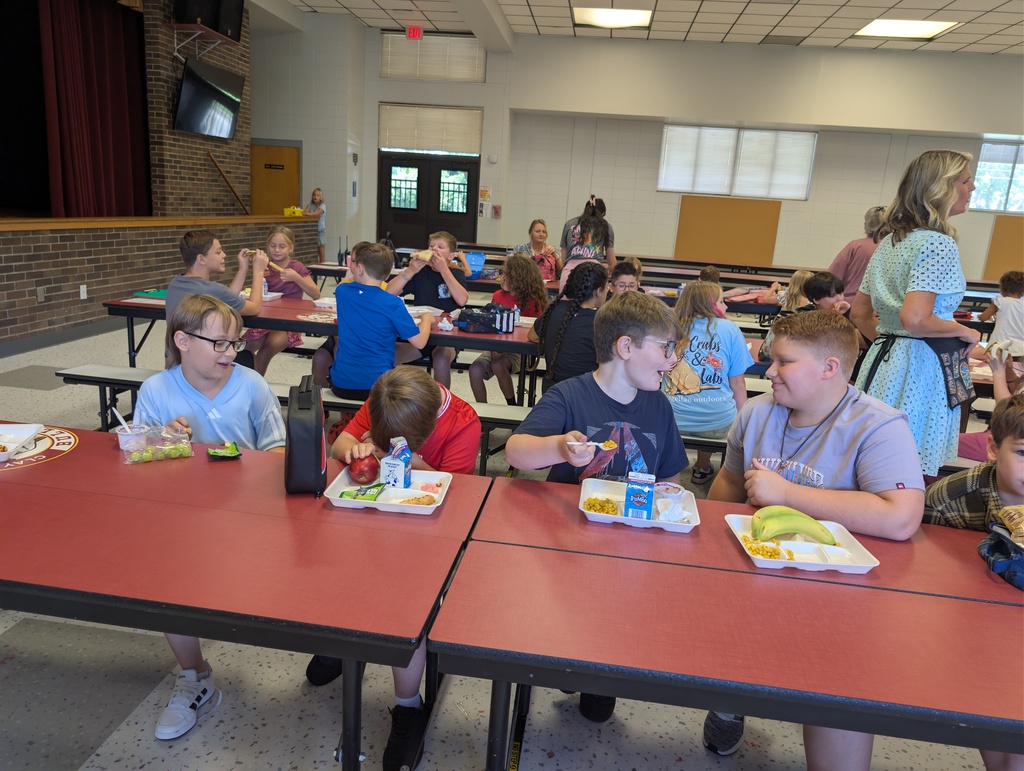 Multiple children eating in a classroom with red tables, some wearing glasses. A woman stands behind them.