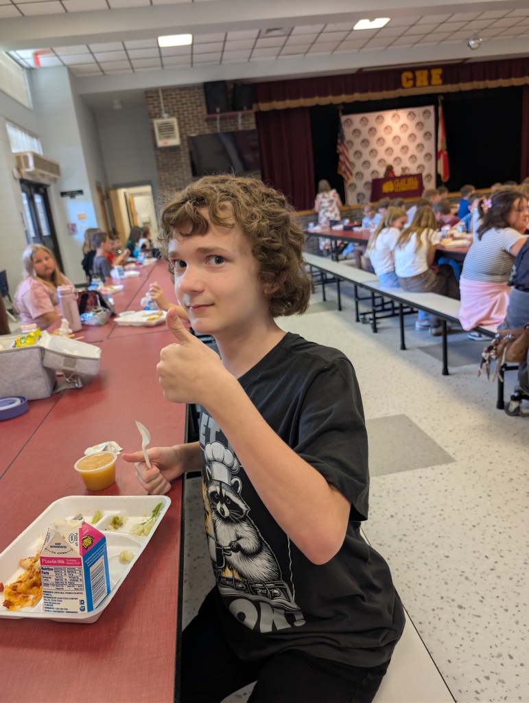 A boy in a black t-shirt sits at a cafeteria table, holding food and giving a thumbs-up.