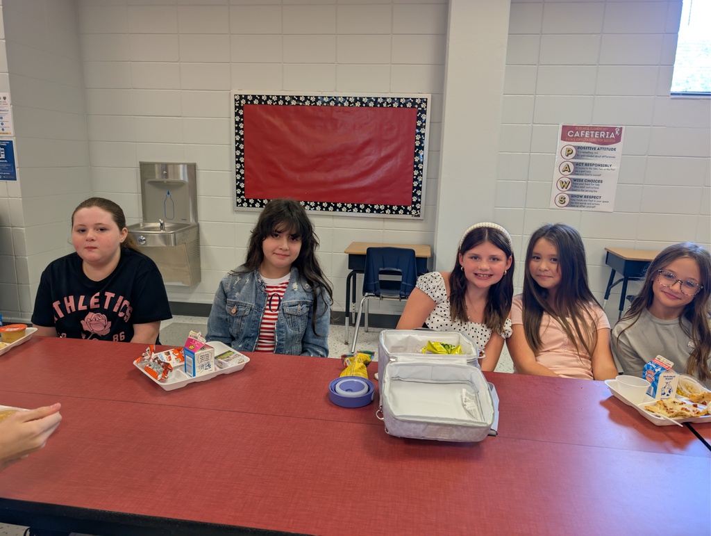 Several children are seated at a table with lunch trays. The table is red. The background has a red bulletin board.