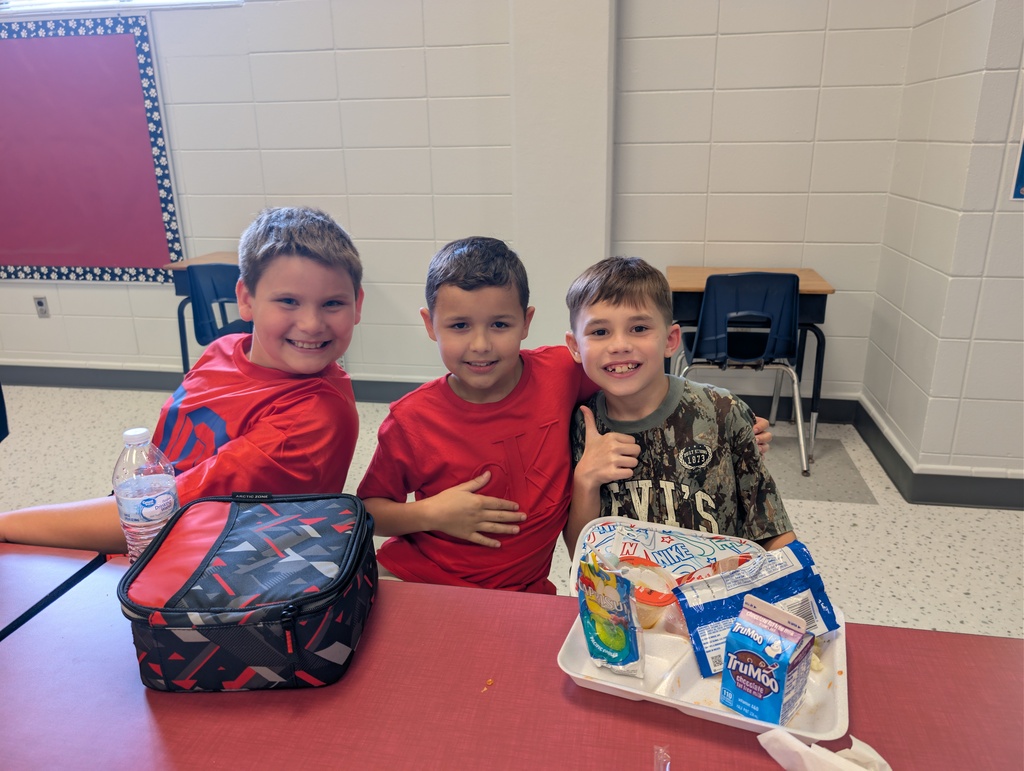 Three boys sit at a table. A lunchbox, a tray of food, and a water bottle are on the table.