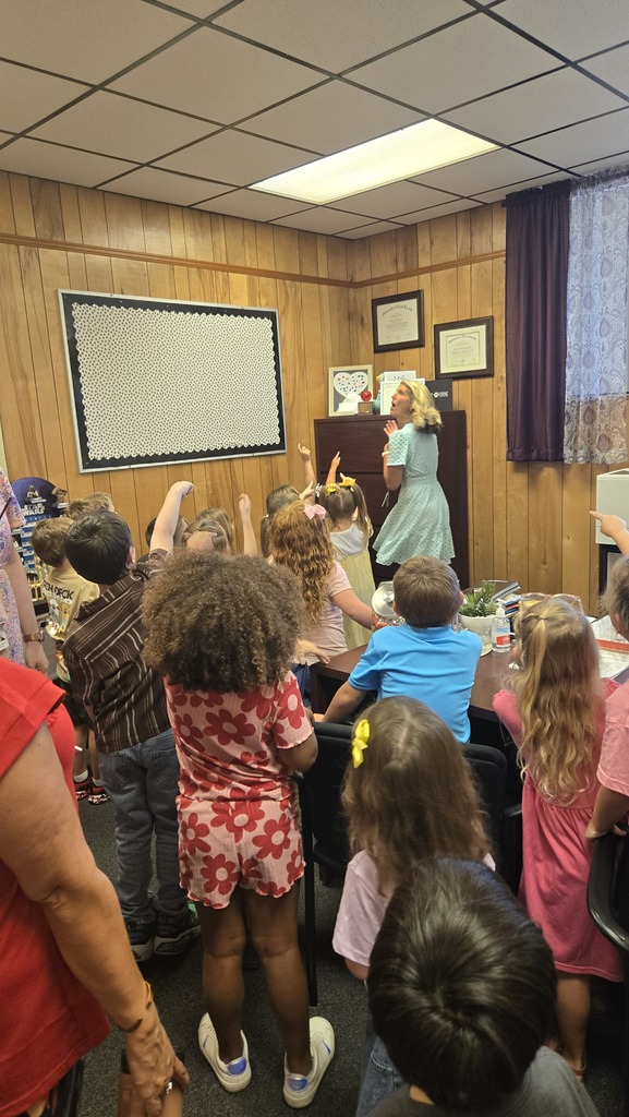 A group of children with curly hair stand in a room with wooden walls. An adult stands at a podium.