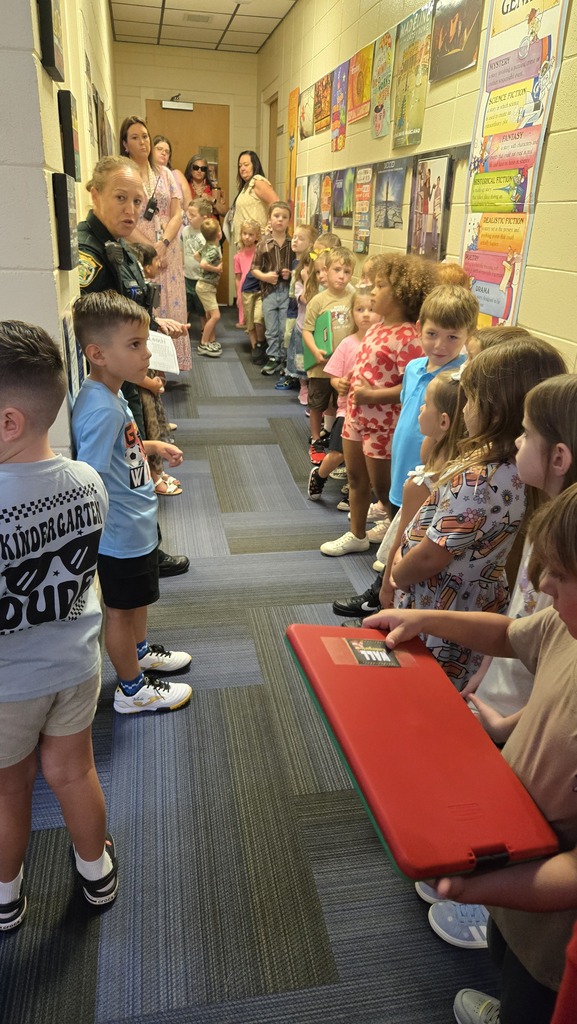 A hallway with children in line. Some wear sneakers. A person holds a red folder.