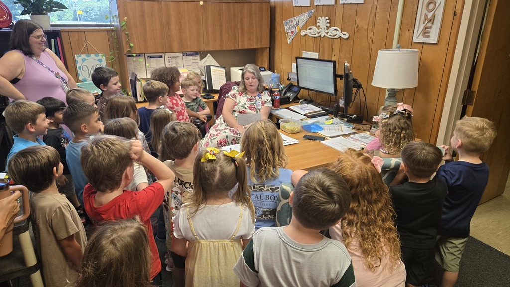 A teacher speaks to students in a classroom. Many students are raising their hands.