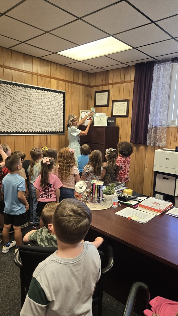 A room with wooden walls has a woman reading to a group of children standing and sitting.