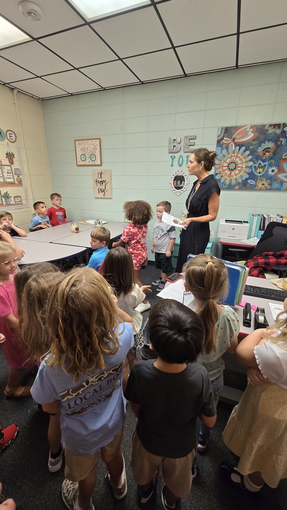 A teacher stands at the front of a classroom with young children, engaged in a lesson.