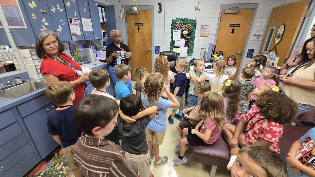 A woman stands at the front of a classroom with children seated and standing around.