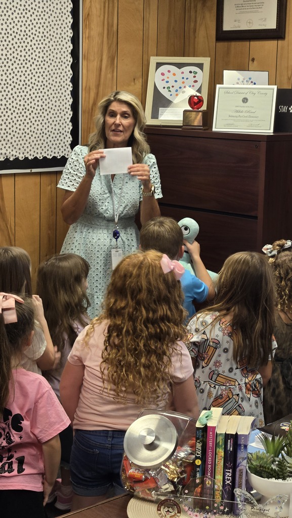 A woman stands in front of children, holding a piece of paper, with a wooden cabinet, bulletin board, and framed picture behind her.