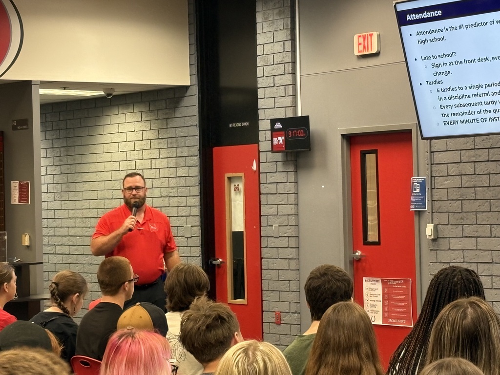Man with microphone stands in front of a red door, speaking to a group of seated students. A monitor with text is visible.
