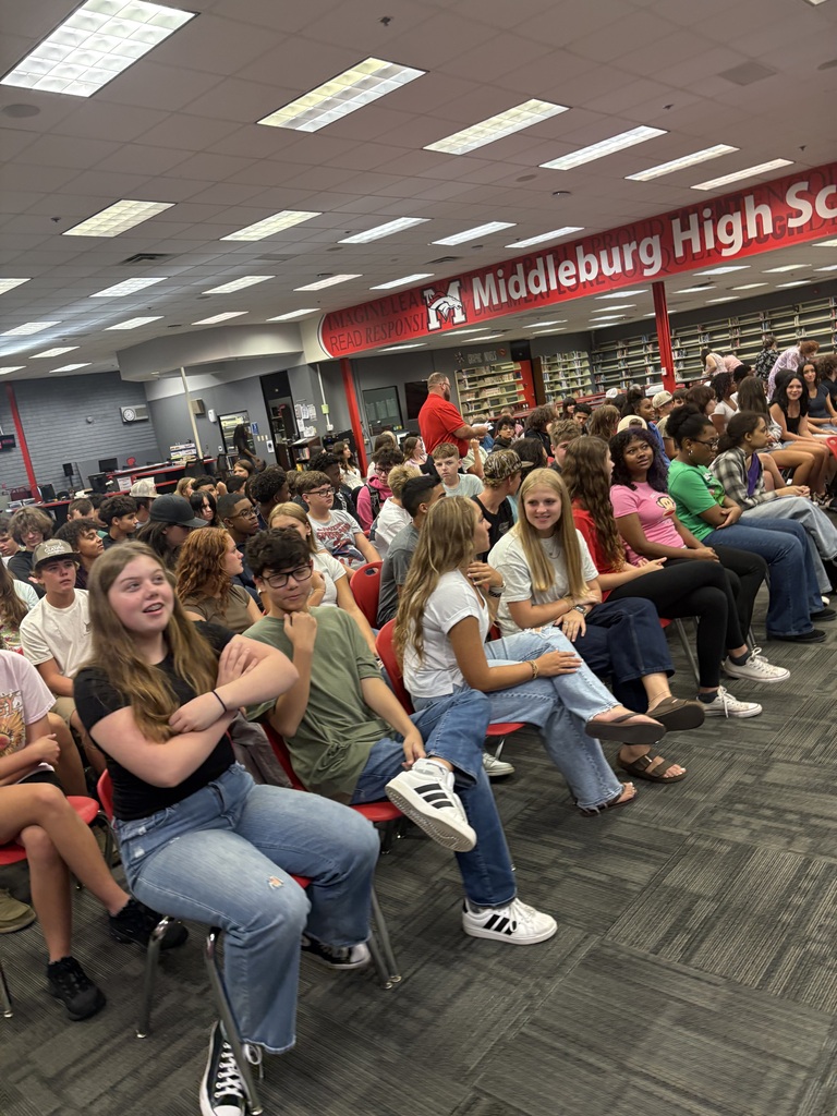 Many people seated in chairs in a school common area, possibly attending an event. The ceiling has lights.