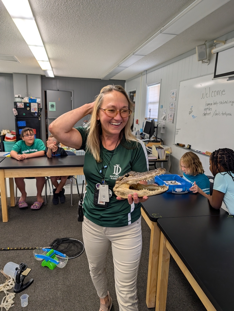 A woman wearing glasses and a green shirt holds an alligator in a classroom. Children sit at desks behind her.