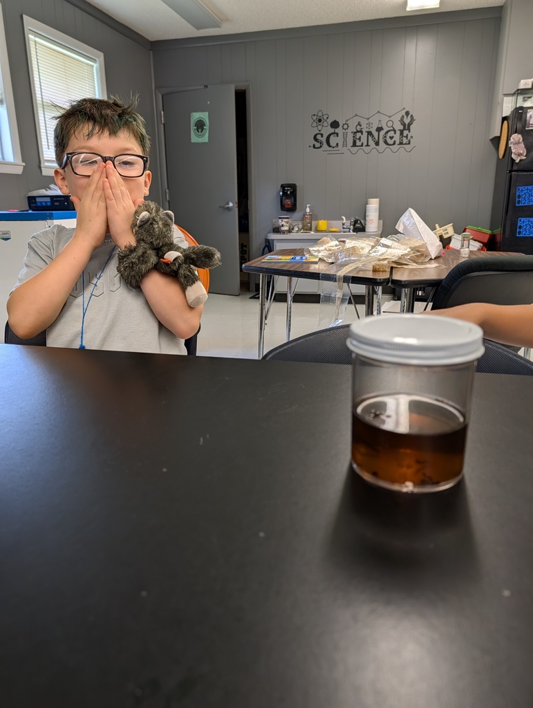 A child in a gray shirt sits at a table with a cup of liquid. The child holds a stuffed animal and has their hands over their face.