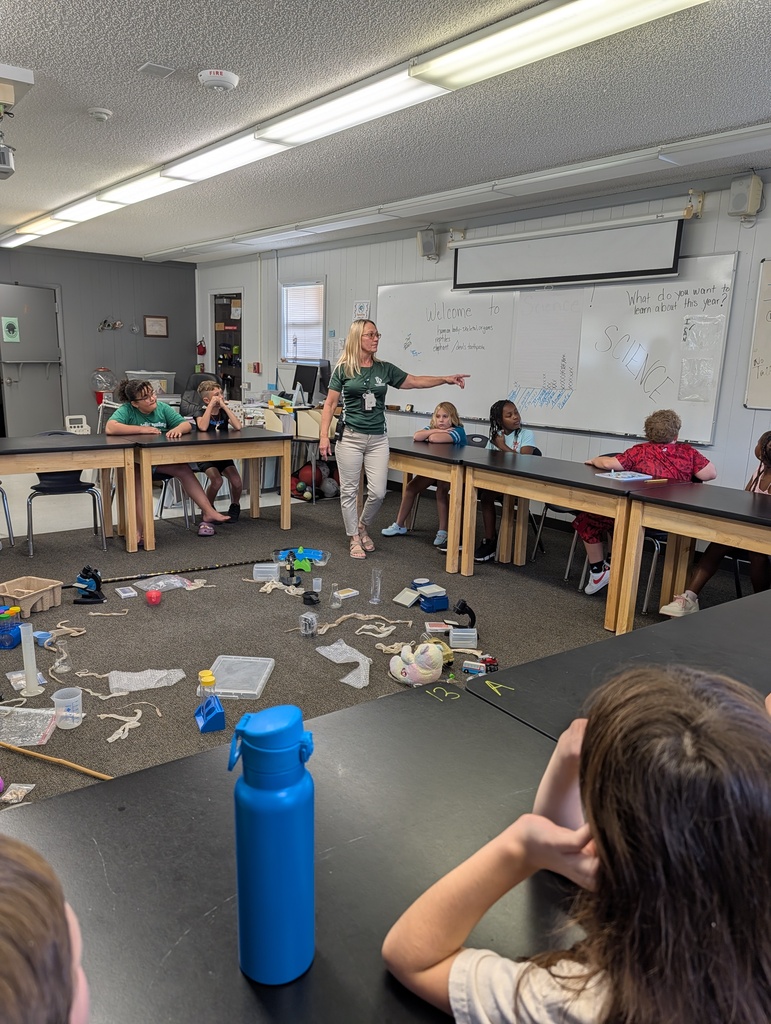 Classroom with tables. A teacher wearing a green shirt and white pants stands at a whiteboard. Students sit at tables, some focused on the teacher. A blue water bottle is on a table.