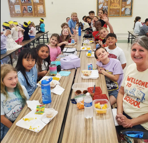 Children seated at tables with food and drinks. A woman wearing a white shirt stands at the end.