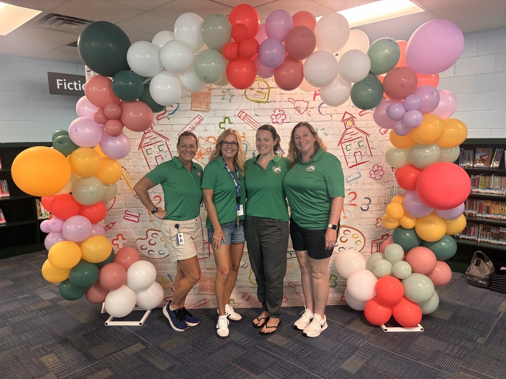 Four people in green shirts and shorts pose under a colorful balloon arch in a library.