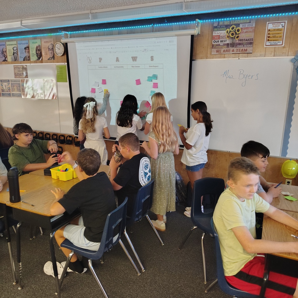 Classroom with children seated at tables, a projector screen displaying information, and a teacher at the front.