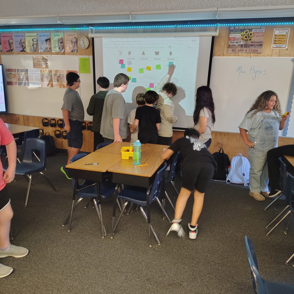 People in a classroom with desks, a whiteboard, and a screen. Some people are standing.