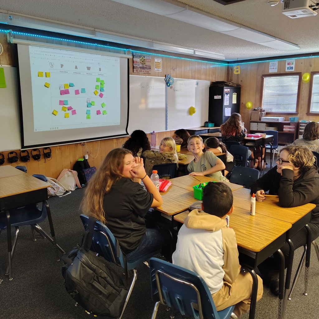 A classroom with tables and chairs, a projector screen on the wall, and people sitting. Some wear coats, and some hold cups.