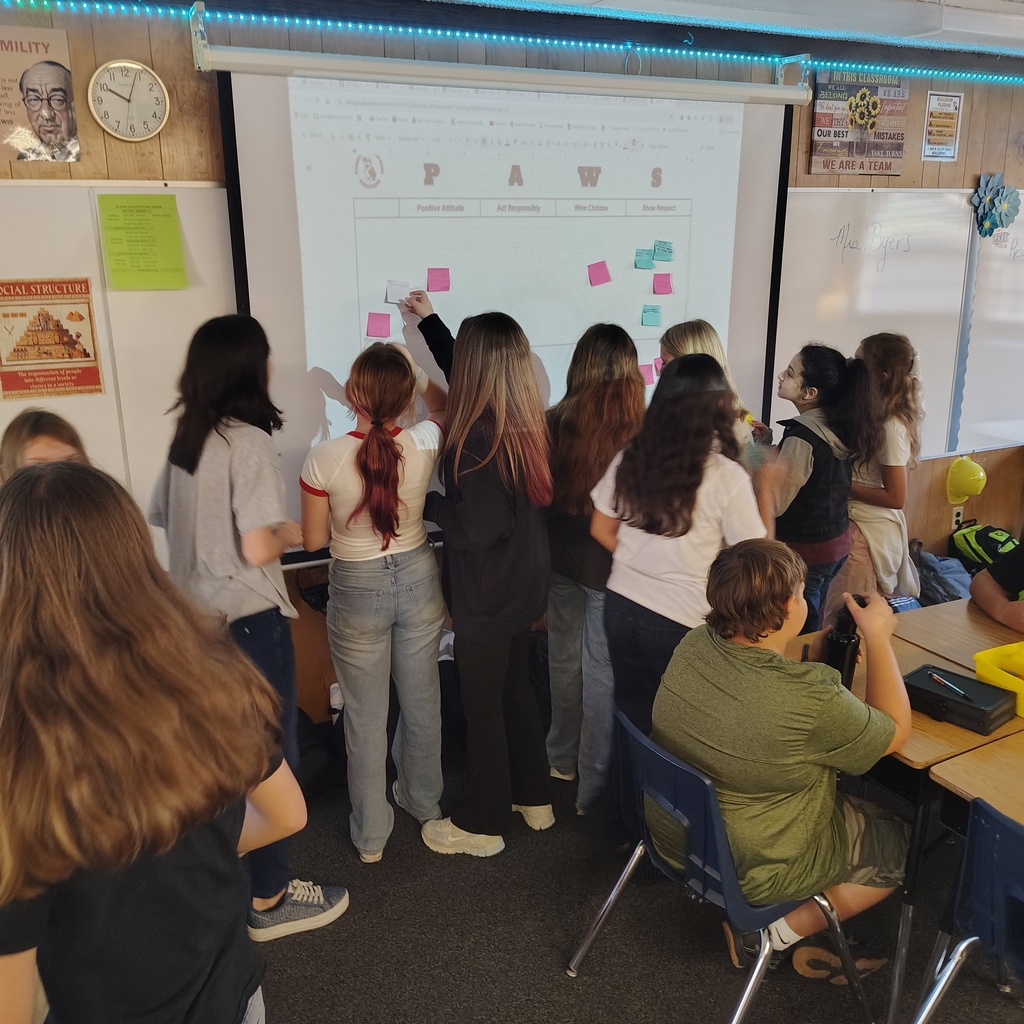 A classroom with people, some standing, some seated, and a teacher using a projector screen with a white board.