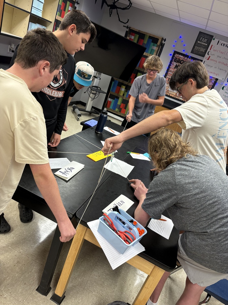 Five students work around a table, examining papers and items in a classroom with bookshelves in the background.