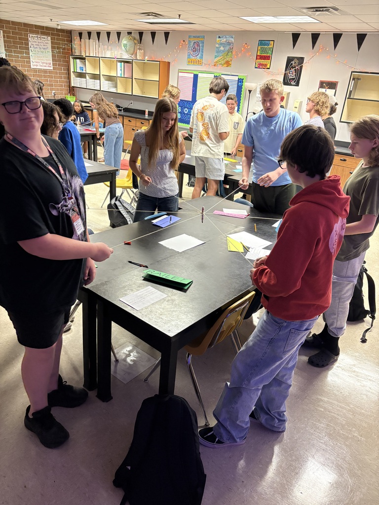 A group of students are gathered around a table in a classroom, engaging in a hands-on activity.