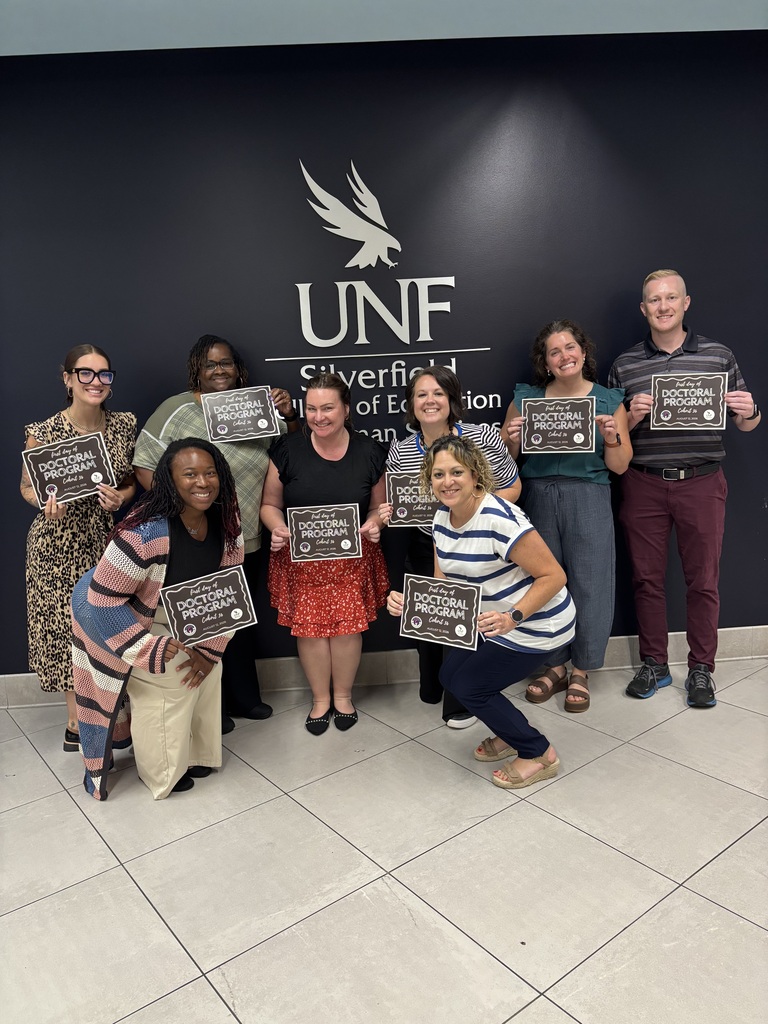 A group of people stand in front of a black wall with the UNF logo, holding certificates.