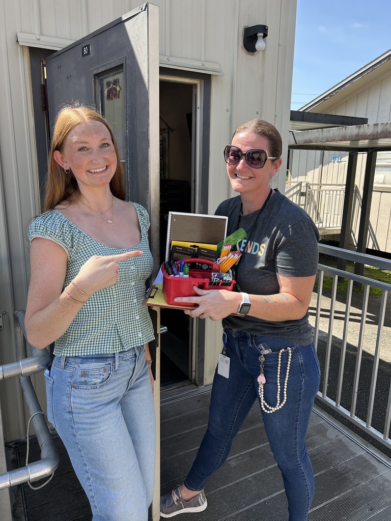 Two women in front of a building. One holds a box with various items, pointing. The other wears a watch and sunglasses.