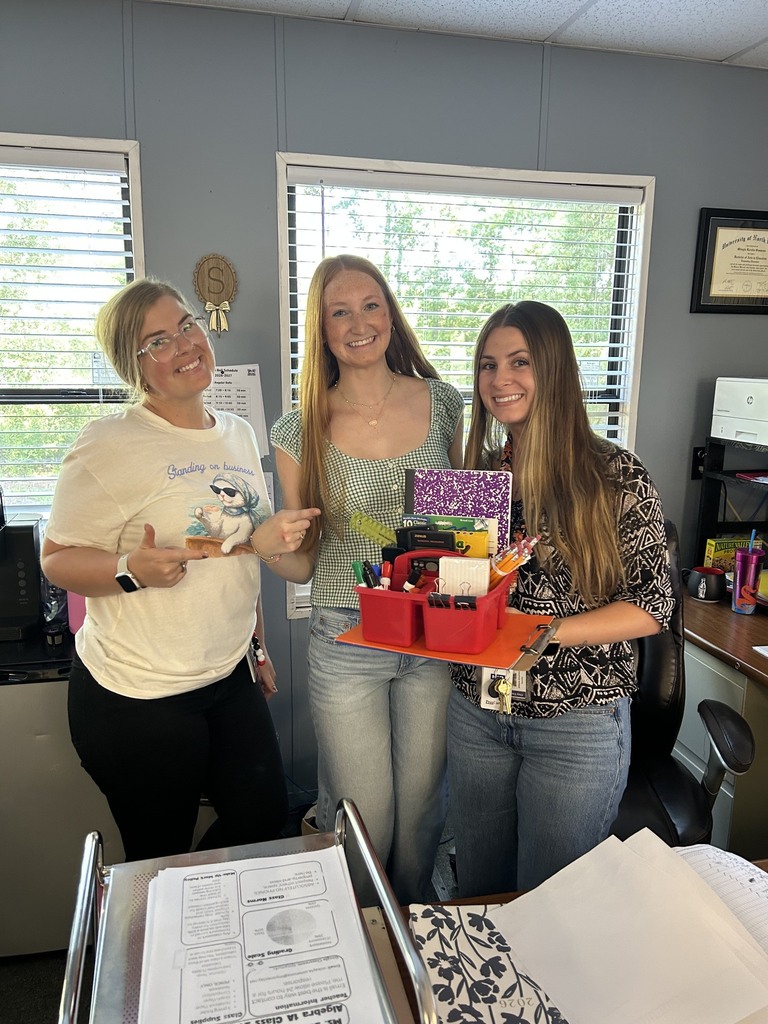 Three women standing in an office. Two hold a basket of school supplies. One gestures with her hand.
