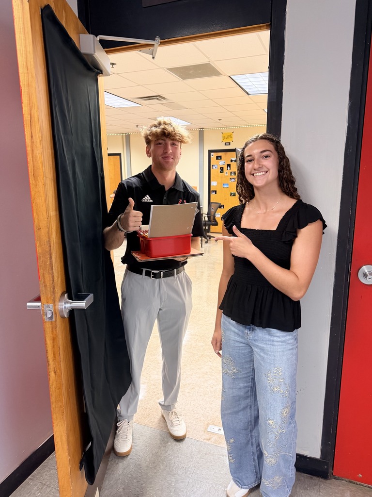 Two people stand at an open door. A man holds a box, and a woman gestures. Both wear casual attire.