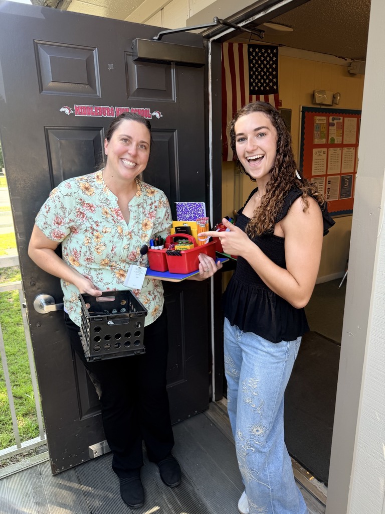 Two women smile in doorway, one holding a box of food. One holds a basket, the other holds a box.