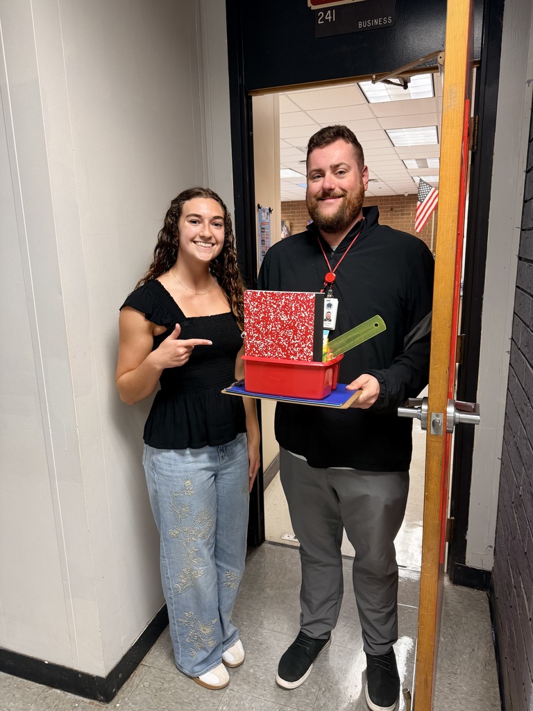Two people pose for a photo. A man in a black jacket holds a red box and a book. A woman in a black shirt smiles.