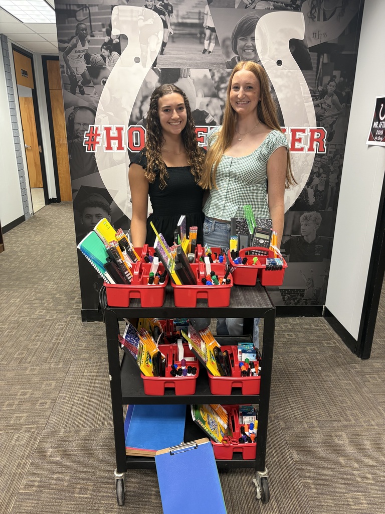 Two women stand next to a cart with trays of supplies and a blue folder. Behind them is a wall with a large horseshoe design and text.