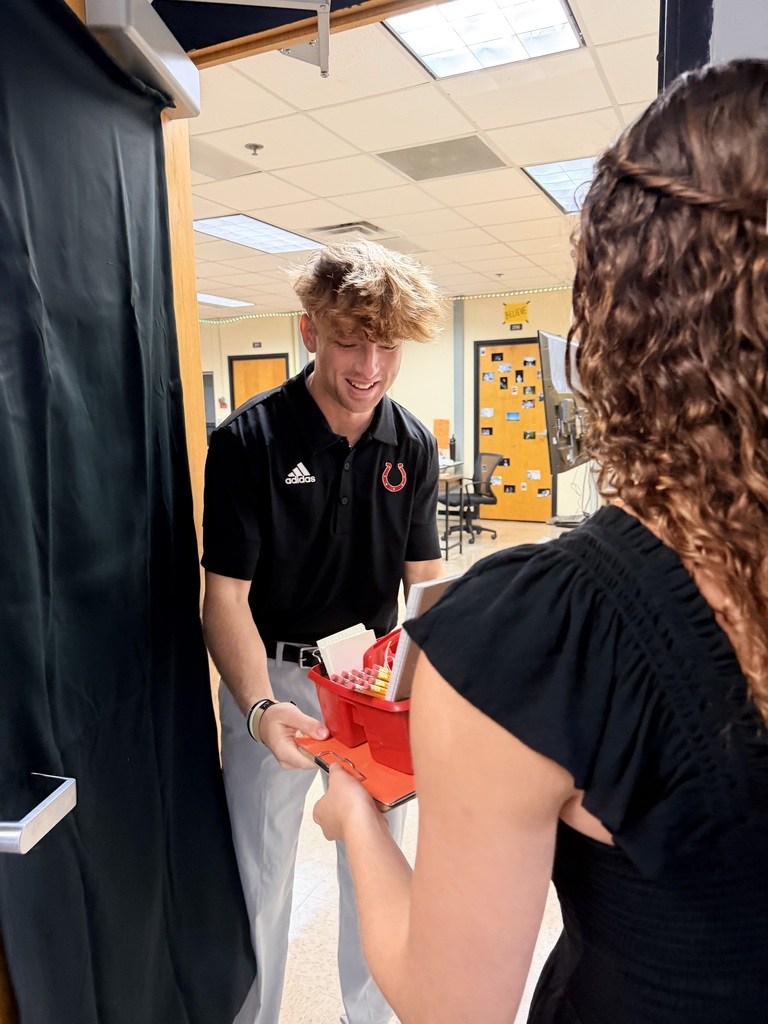A man in a black shirt presents a red box to a woman with curly hair in a hallway.