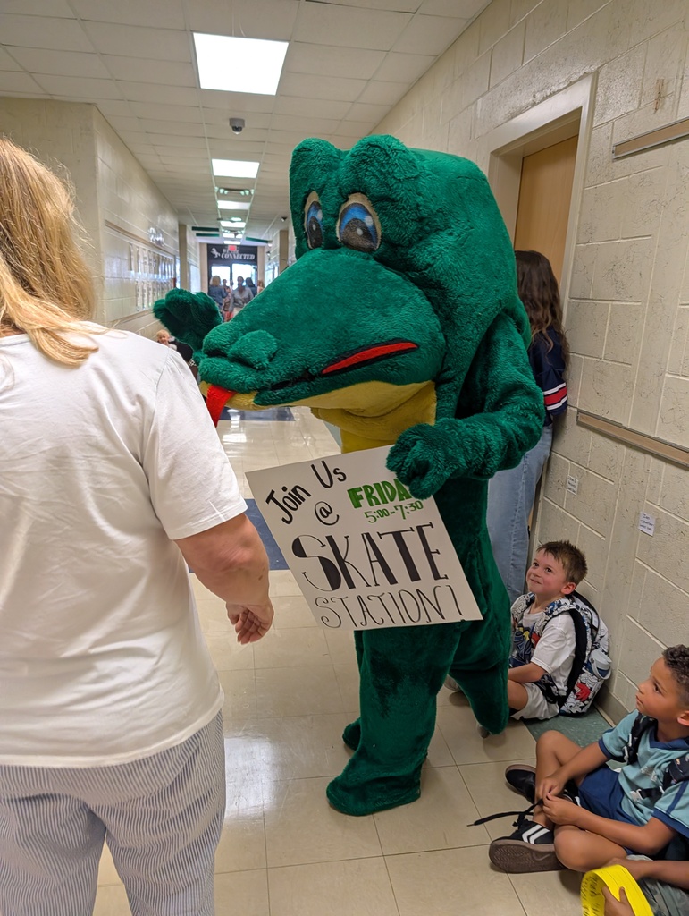 A mascot dressed as a crocodile holds a sign while a woman looks on. Behind, two children sit on the floor.