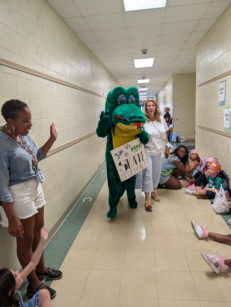A mascot, dressed in green, walks through a school hallway with a sign. A woman in white shorts stands nearby. Students sit on the floor in the background.