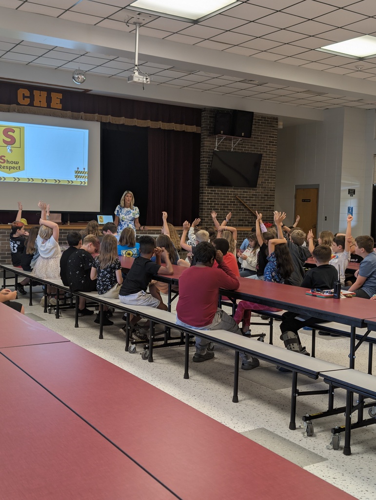 A classroom with rows of students seated at tables, a teacher standing in front of a projector screen.
